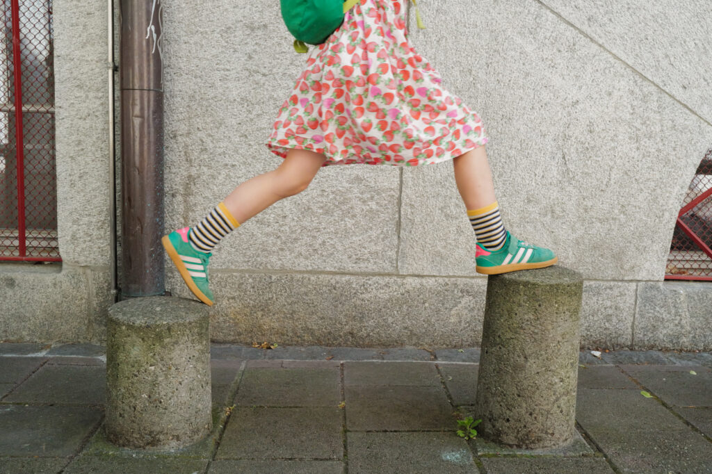 Child in a strawberry-print dress leaping between two stone bollards, green sneakers, Basel.