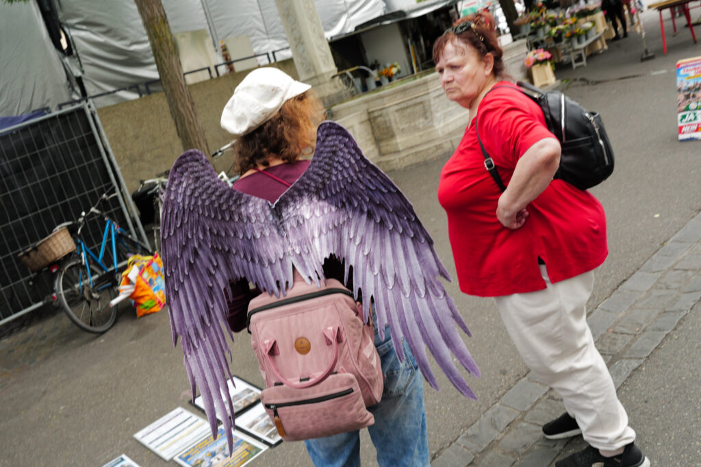 Person wearing large purple angel wings and a pink backpack at a market, a woman in red looking at them, Basel.