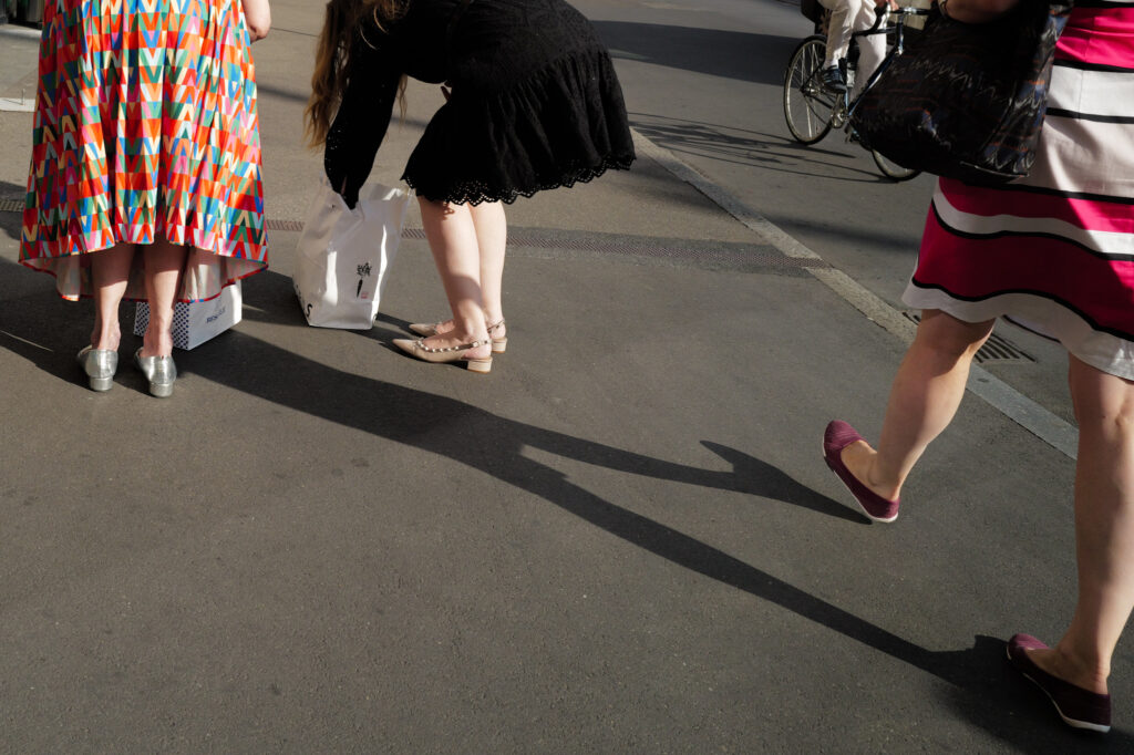 Three pairs of legs and long shadows on pavement, one person carrying a white shopping bag, Basel.