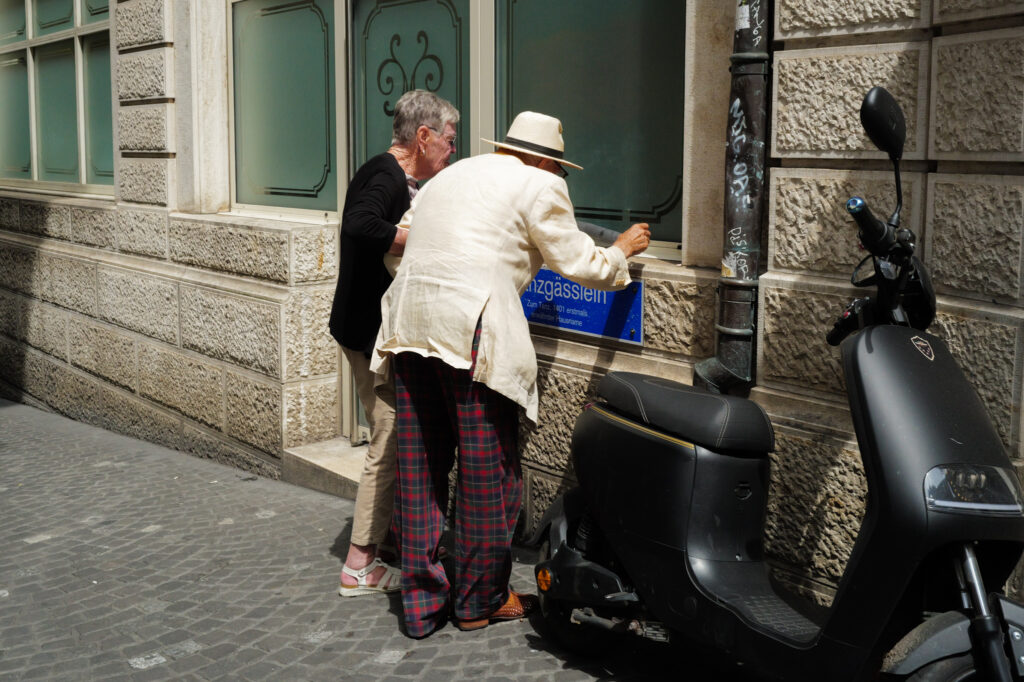 Elderly couple reading a street-name plaque on a stone wall, scooter parked nearby. Basel.