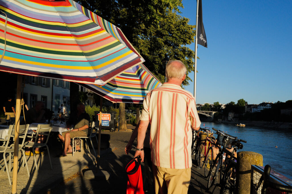 Man in striped shirt carrying a red bag walking along the Rhine promenade in Basel, colorful umbrellas.