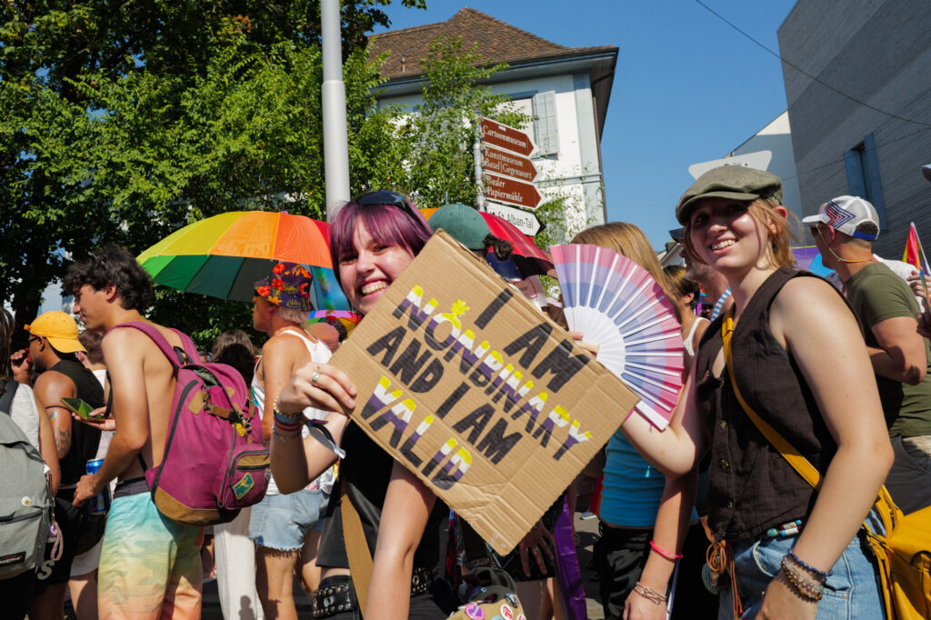 Smiling people at Basel Pride, one holding a cardboard sign reading “I am nonbinary and I am valid,” with rainbow umbrellas and a Basel street sign behind.