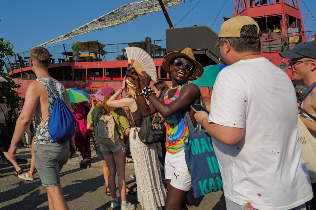 A man in a rainbow tank top and straw hat laughs while raising a paper fan at Basel Pride, a red ship-restaurant behind him in the crowd.