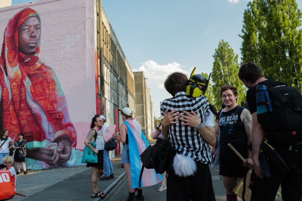 Two people embrace at a Pride gathering in Basel, one wearing a yellow-and-black pup mask, in front of a large mural of a woman in a red headscarf; people with trans flags stand nearby along an industrial street.