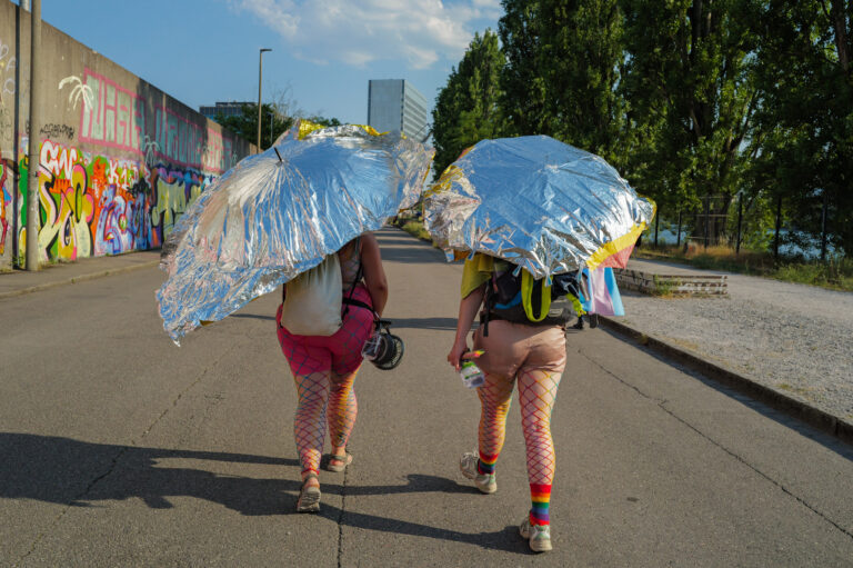 Two figures seen from behind walk along a Basel street during Pride, shaded by umbrellas wrapped in silver thermal blankets, wearing rainbow fishnet tights and carrying backpacks and a trans flag.
