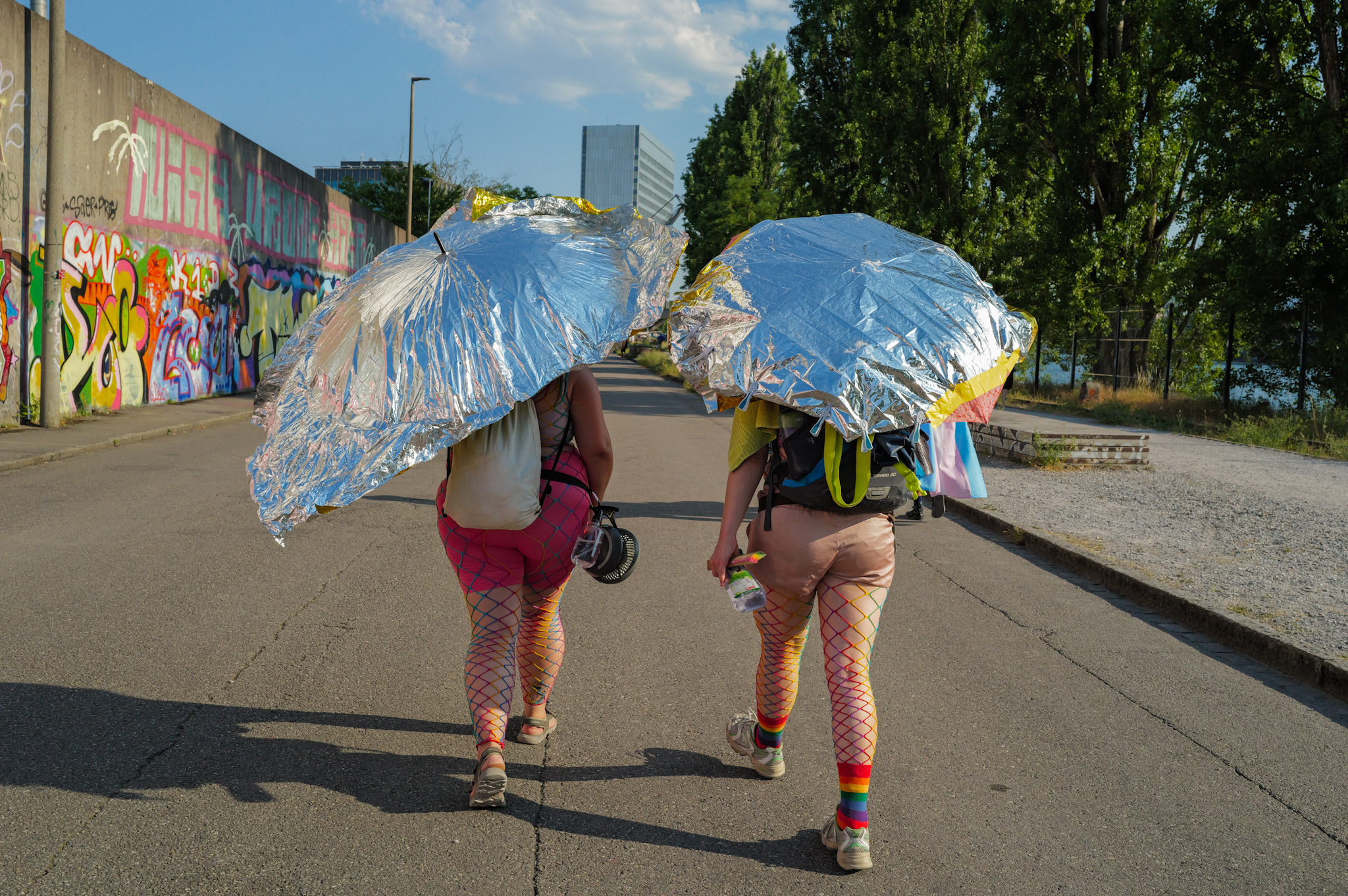 Two figures seen from behind walk along a Basel street during Pride, shaded by umbrellas wrapped in silver thermal blankets, wearing rainbow fishnet tights and carrying backpacks and a trans flag.